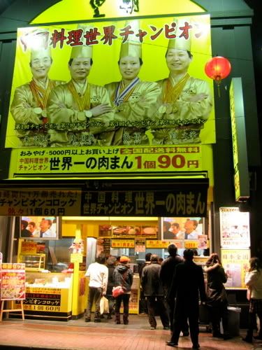 「世界一の肉まん」看板に料理人4人の写真、その下で列をなす買物客